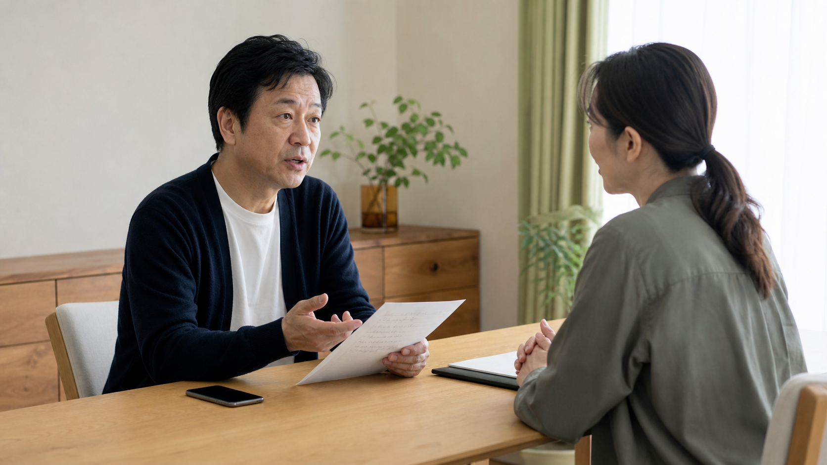 相談メモを手にした男性が専門職と対話し、机上には閉じたスマートフォンが置かれている